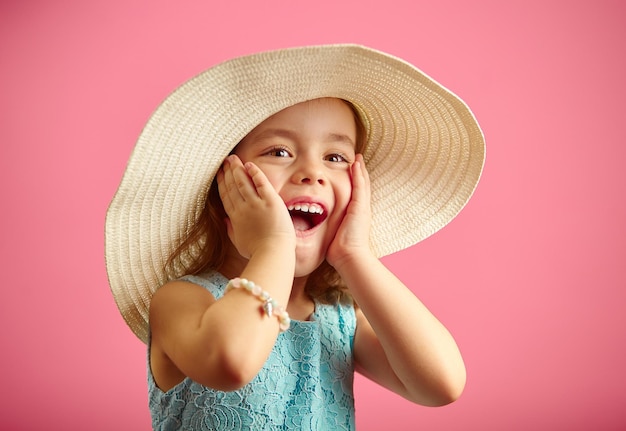 Petite fille surprise en chapeau de paille et robe bleue met ses mains face à face exprime sa surprise