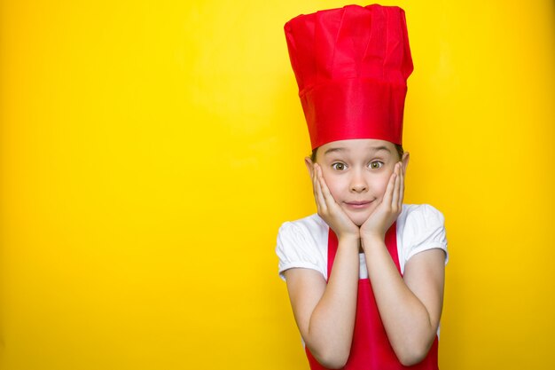 Petite fille surprise en costume de chef rouge avec les mains sur les joues sur jaune