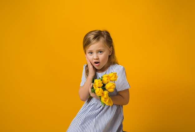 Une petite fille surprise dans une robe rayée est assise et tient un bouquet de tulipes jaunes sur jaune