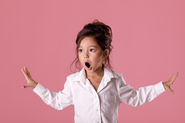 Petite fille surprise mignonne en chemise blanche avec coiffure à la recherche d'appareil photo sur fond rose. Émotions humaines et expression faciale