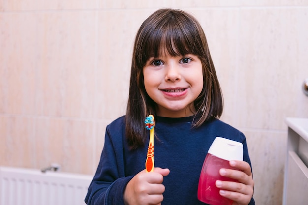 Petite fille en sweat bleu foncé, souriante, tenant une brosse à dents et du dentifrice, dans la salle de bain. Concept de brossage des dents, d'hygiène et d'enfance.