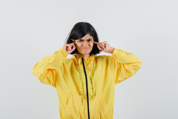 Photo petite fille en sweat à capuche jaune, brancher les oreilles avec les doigts et à la vue de face, irrité.