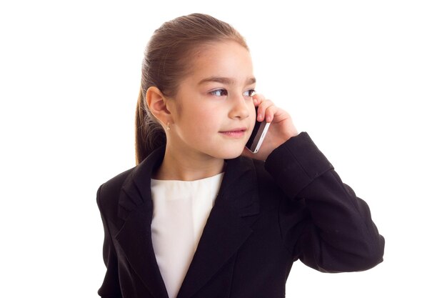 Petite fille sympa avec une longue queue de cheval marron en T-shirt blanc, veste noire parlant au téléphone sur fond blanc en studio