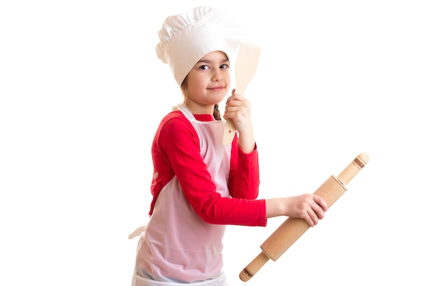 Petite fille sympa tenant un rouleau à pâtisserie et une spatule de cuisine sur fond blanc en studio