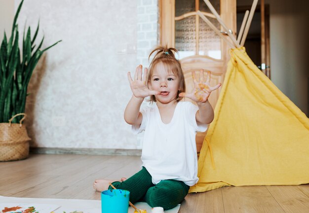 Une petite fille en t-shirt blanc peint des maisons sur le sol et montre ses mains sales. Jeux pour enfants