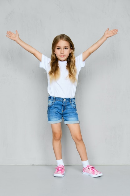 Petite fille en t-shirt blanc, short en jean et baskets roses debout avec les mains levées près du mur