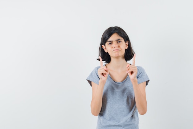 Petite fille en t-shirt pointant vers le haut et regardant sans espoir, vue de face.