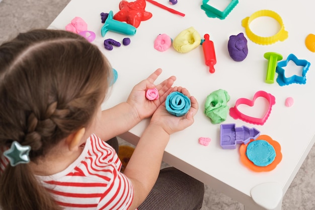 Petite fille en T-shirt à rayures lumineuses jouant avec de la pâte à modeler faisant de la pâte à modeler de fleur rose sur le concept d'artisanat d'art de table blanche