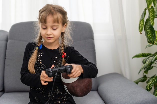 Petite fille tenant un ballon de rugby et un joystick pour jouer à des jeux vidéo