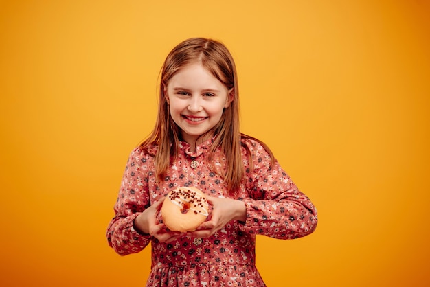 Petite fille tenant un beignet dans les mains et regardant la caméra en souriant isolée sur fond jaune avec fond. Jolie fille avec des bonbons pendant la séance photo en studio