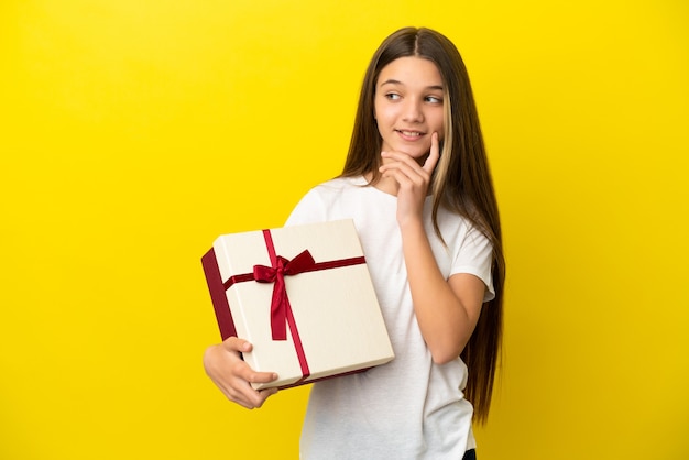 Petite fille tenant un cadeau sur fond jaune isolé pensant à une idée tout en levant les yeux