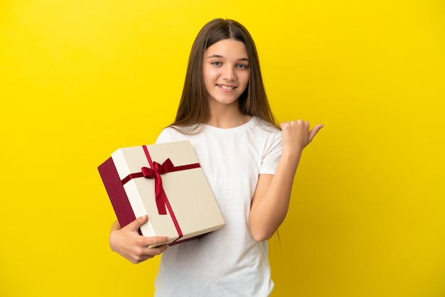 Petite fille tenant un cadeau sur fond jaune isolé pointant vers le côté pour présenter un produit