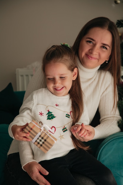 Petite fille tenant un cadeau et serrant sa maman devant l'arbre du Nouvel An