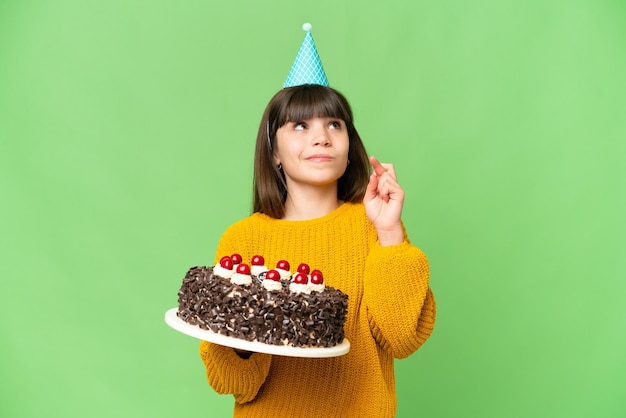 Petite fille tenant un gâteau d'anniversaire sur fond de clé chroma isolé avec les doigts croisés et souhaitant le meilleur