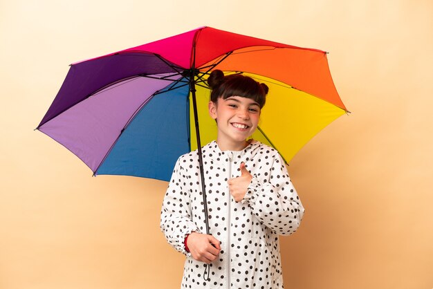 Petite fille tenant un parapluie isolé sur beige donnant un geste de pouce en l'air