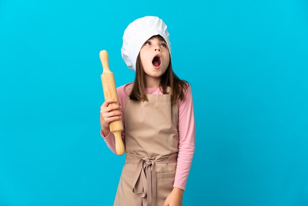 Petite fille tenant un rouleau à pâtisserie isolé sur fond bleu en levant et avec une expression de surprise