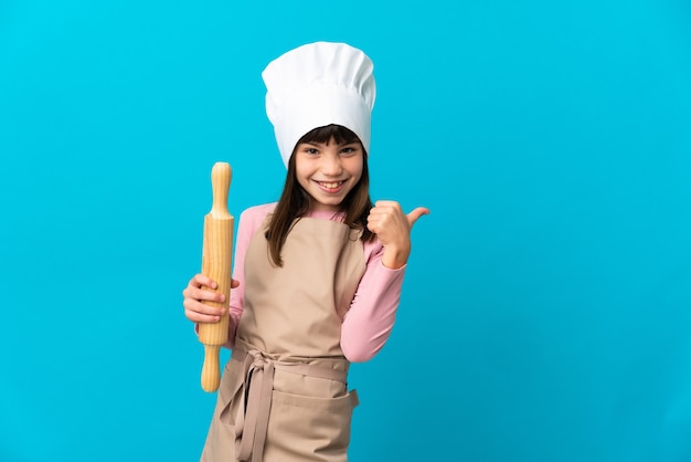 Petite fille tenant un rouleau à pâtisserie isolé sur un mur bleu pointant vers le côté pour présenter un produit
