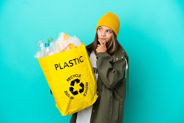 Petite fille tenant un sac plein de bouteilles en plastique à recycler sur fond bleu isolé ayant des doutes