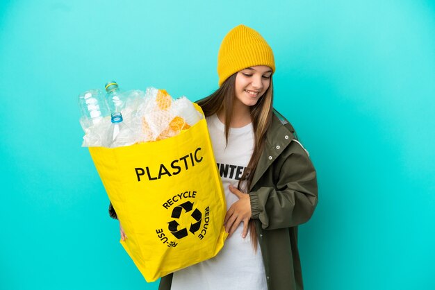 Petite fille tenant un sac plein de bouteilles en plastique à recycler sur un mur bleu isolé souriant beaucoup