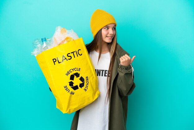 Petite fille tenant un sac plein de bouteilles en plastique à recycler sur une surface bleue isolée pointant vers le côté pour présenter un produit