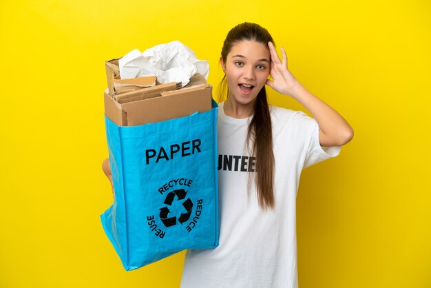 Petite fille tenant un sac de recyclage plein de papier à recycler sur fond jaune isolé avec expression surprise