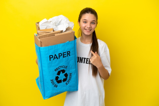 Petite fille tenant un sac de recyclage plein de papier à recycler sur une surface jaune isolée avec une expression faciale surprise