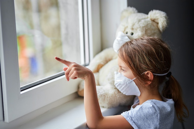 Petite fille tenant et serrant l'ours en peluche dans un masque près de la fenêtre.