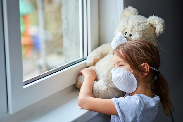 Petite fille tenant et serrant l'ours en peluche dans un masque près de la fenêtre.