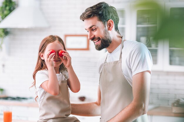 Petite fille tenant des tomates cerises rouges sur les yeux avec le père debout près de la cuisine