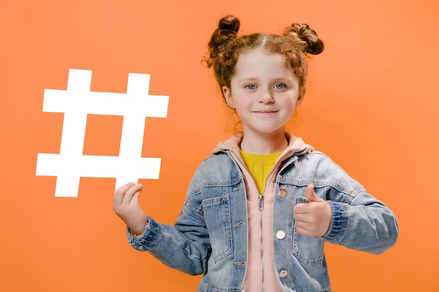 petite fille tenir l'icône de hashtag blanc et avec son pouce vers le haut isolé sur fond orange