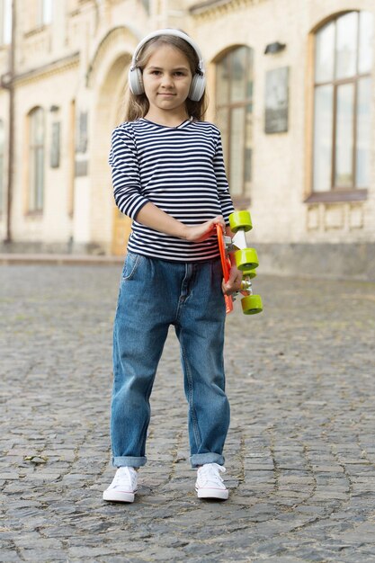 Petite fille tenir penny board en écoutant de la musique dans des écouteurs en plein air urbain d'été, en voyage.