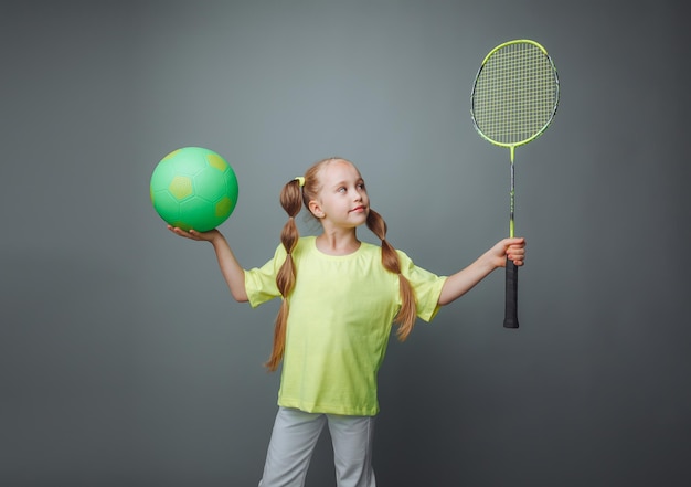 Une petite fille tient une balle et une raquette sur un fond gris