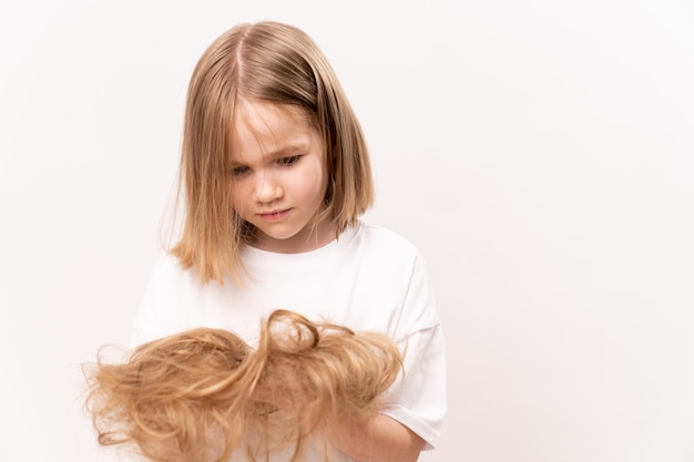 Une petite fille tient dans les mains les cheveux coupés après avoir coupé sur un fond blanc. signifie prendre soin des cheveux des enfants. Salon de beauté pour enfants.