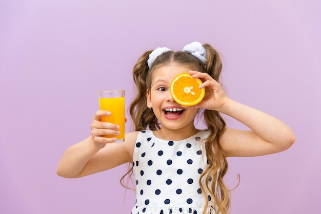 Une petite fille tient du jus d'orange et une orange Un enfant vêtu d'une robe à pois boit du jus fraîchement pressé