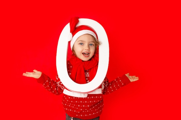 Une petite fille tient le numéro zéro dans le chapeau du père Noël sur fond rouge, espace pour le texte