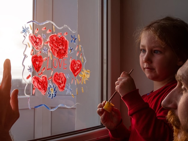 Petite fille tient un pinceau à la main dessinant un coeur rouge sur la vitre Saint Valentin amour rencontres loisirs de quarantaine