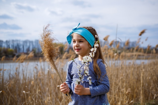 Une petite fille tient des roseaux secs et une branche avec de petites fleurs blanches dans les mains, un printemps ensoleillé, le sourire et la joie de l'enfant