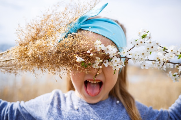 Une petite fille tient des roseaux secs et une branche avec de petites fleurs blanches dans les mains, un printemps ensoleillé, le sourire et la joie de l'enfant