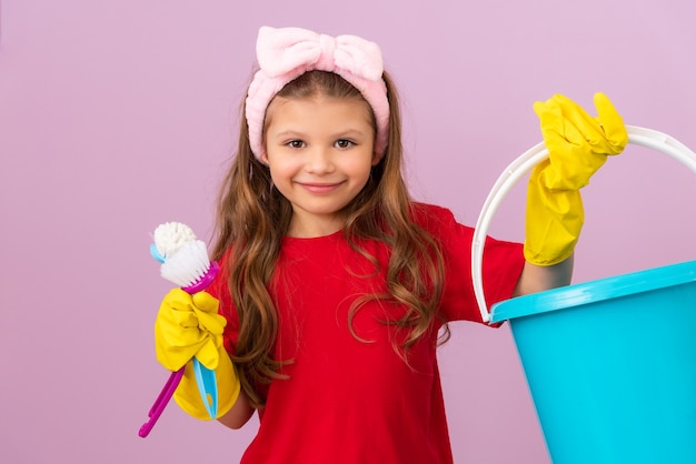 Une petite fille tient un seau en plastique et des brosses pour nettoyer la pièce.