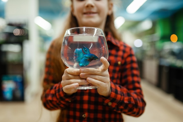 Petite fille tient un verre avec du poisson bleu, animalerie. Enfant achetant du matériel en animalerie, accessoires pour animaux domestiques