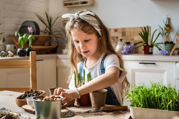 Une petite fille transplante des fleurs et des plantes d'intérieur un enfant dans un bandana plante des bulbes et des micro-verts