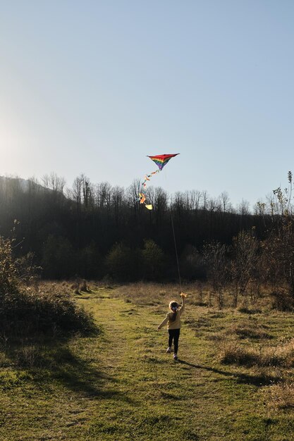 Petite fille traverse le champ sur l'herbe verte au coucher du soleil et lance un cerf-volant multicolore dans le ciel