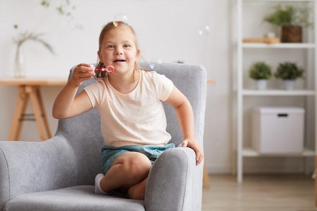 Petite fille trisomique assise sur un fauteuil et faire des bulles à la maison