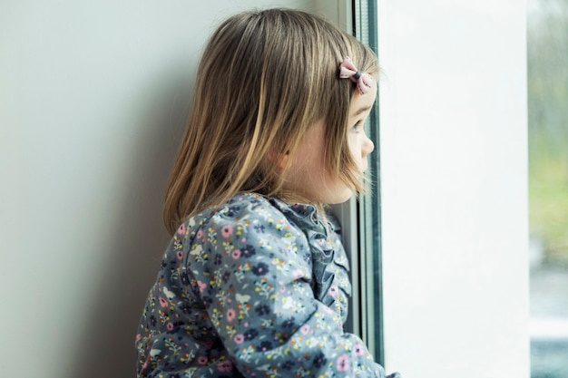 Une petite fille triste regarde par la fenêtre. Enfant de 3 ans en robe. Fermer. La solitude et la tristesse.