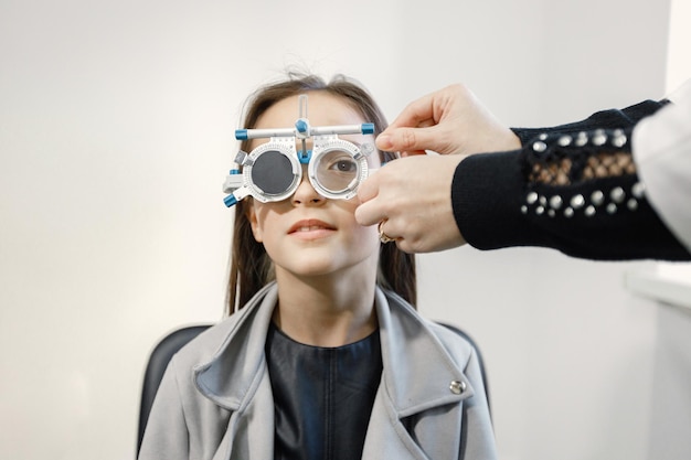 Petite fille vérifiant sa vue au centre d'ophtalmologie. Optométriste féminin faisant des tests de vue. Jeune fille brune portant une veste grise et une chemise noire.