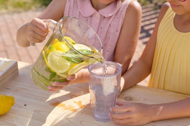 Petite Fille Verser De La Limonade Maison Fraîche Dans Un Verre En Plastique Tout En Vendant Des Boissons Avec Son Amie Par Décrochage Sur Une Chaude Journée D'été