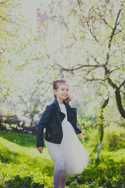 Petite fille en veste noire et robe blanche courant dans le jardin de cerisiers de printemps