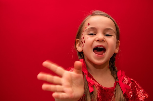 Une petite fille vêtue d'une élégante robe rouge à paillettes sur fond rouge avec des nattes sourit joyeusement en agitant une main graissée à l'appareil photo