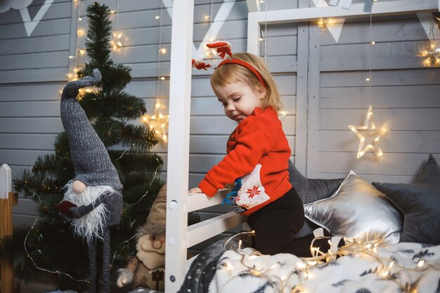 Une petite fille vêtue d'un pull chaud rouge est assise sur un lit près d'un arbre de Noël avec des jouets et des cadeaux. Enfance heureuse. Ambiance de vacances du nouvel an