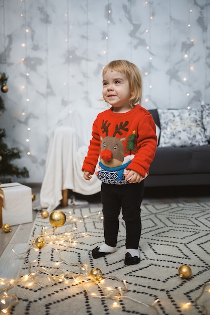 Une petite fille vêtue d'un pull chaud rouge se tient près d'un arbre de Noël avec des jouets et des cadeaux. Enfance heureuse. Ambiance de vacances du nouvel an